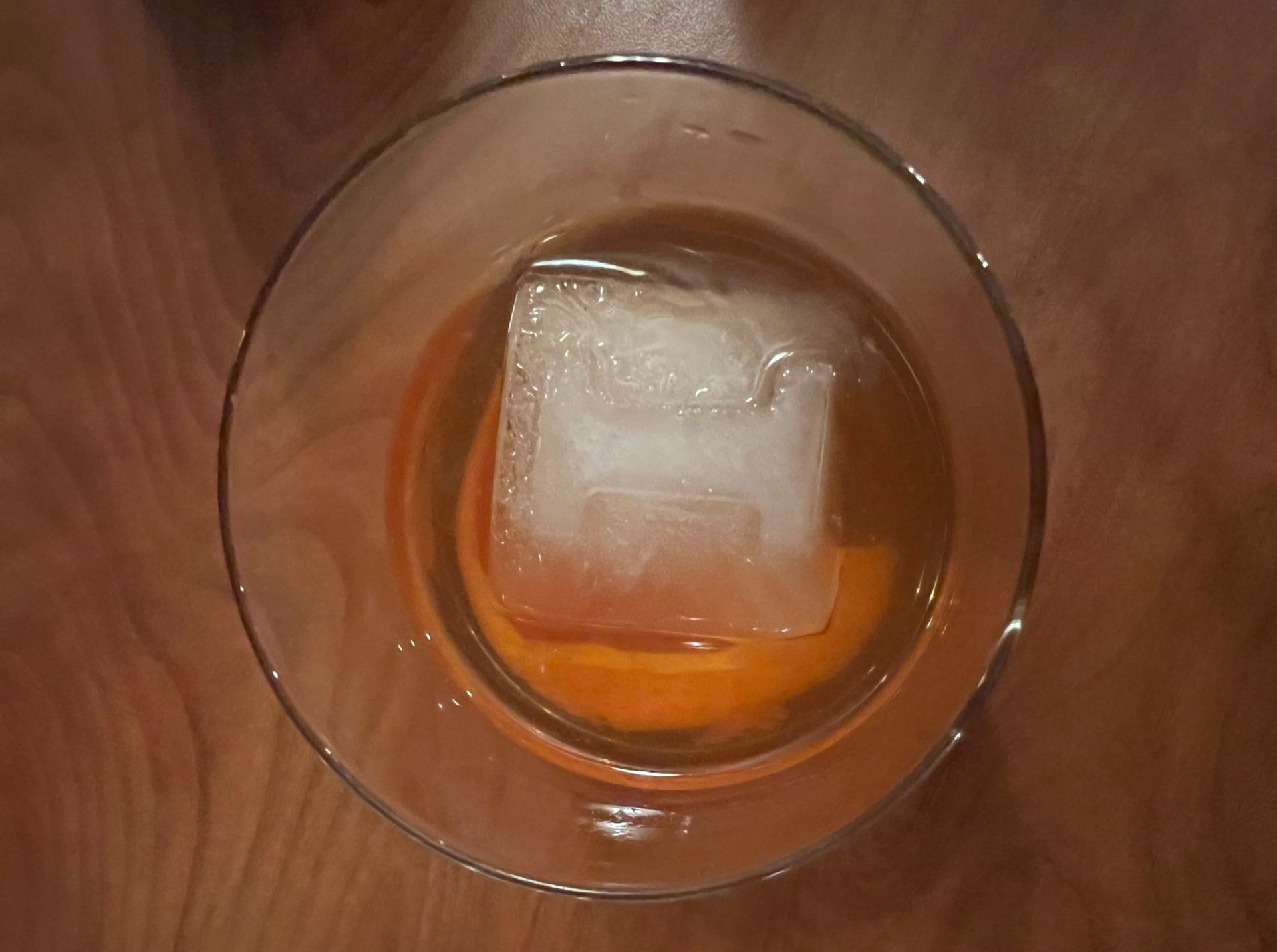 View from above of a clear rocks glass with a light brown liquid in it. There is a large cube of ice with a block "I" carved into it.