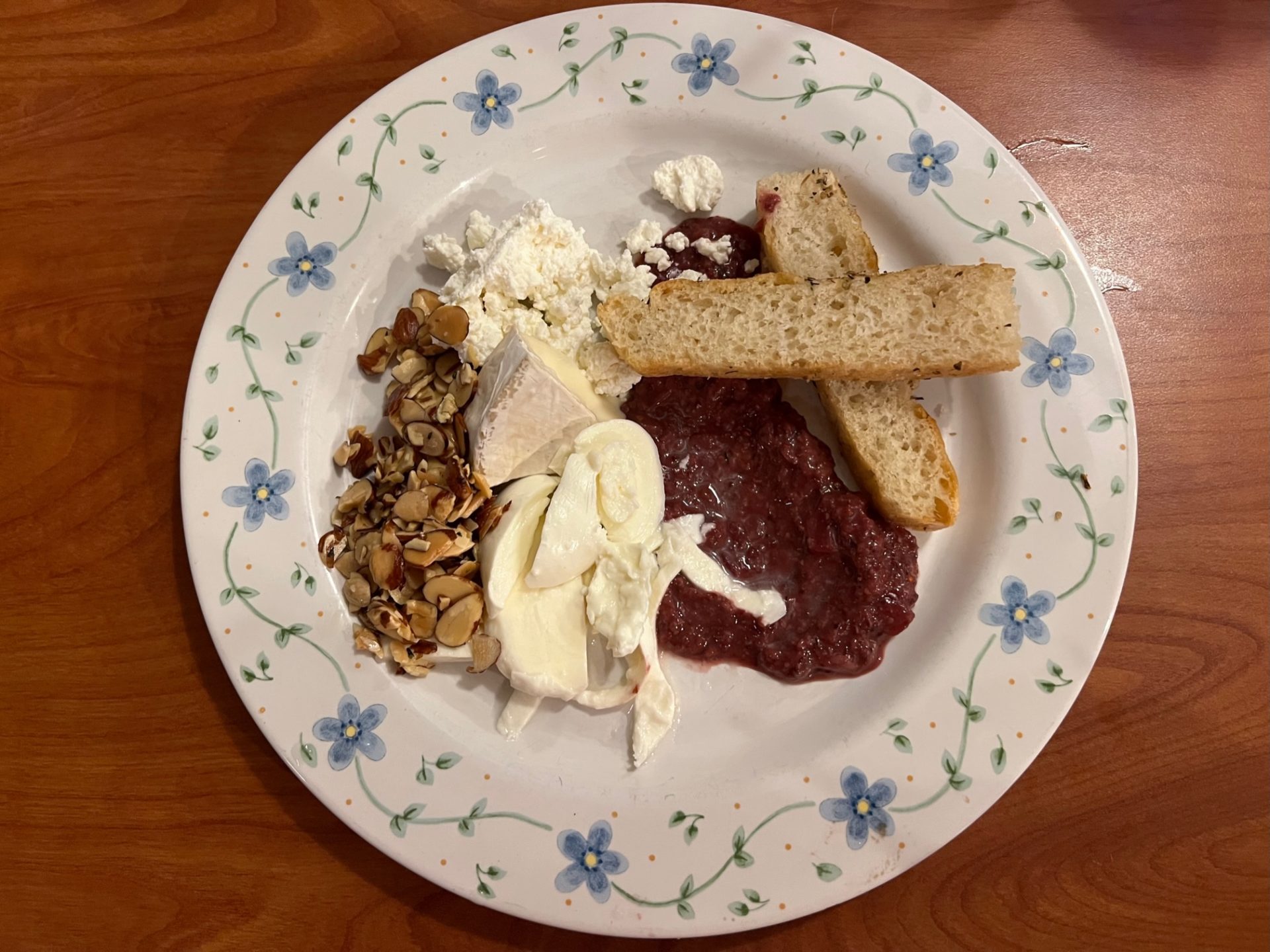 A round white plate with blue flower detailing. On it is a small pile of slivered almonds, three different types of cheese a smear of dark red mostarda, and two small pieces of focaccia criss-crossed on top of each other.