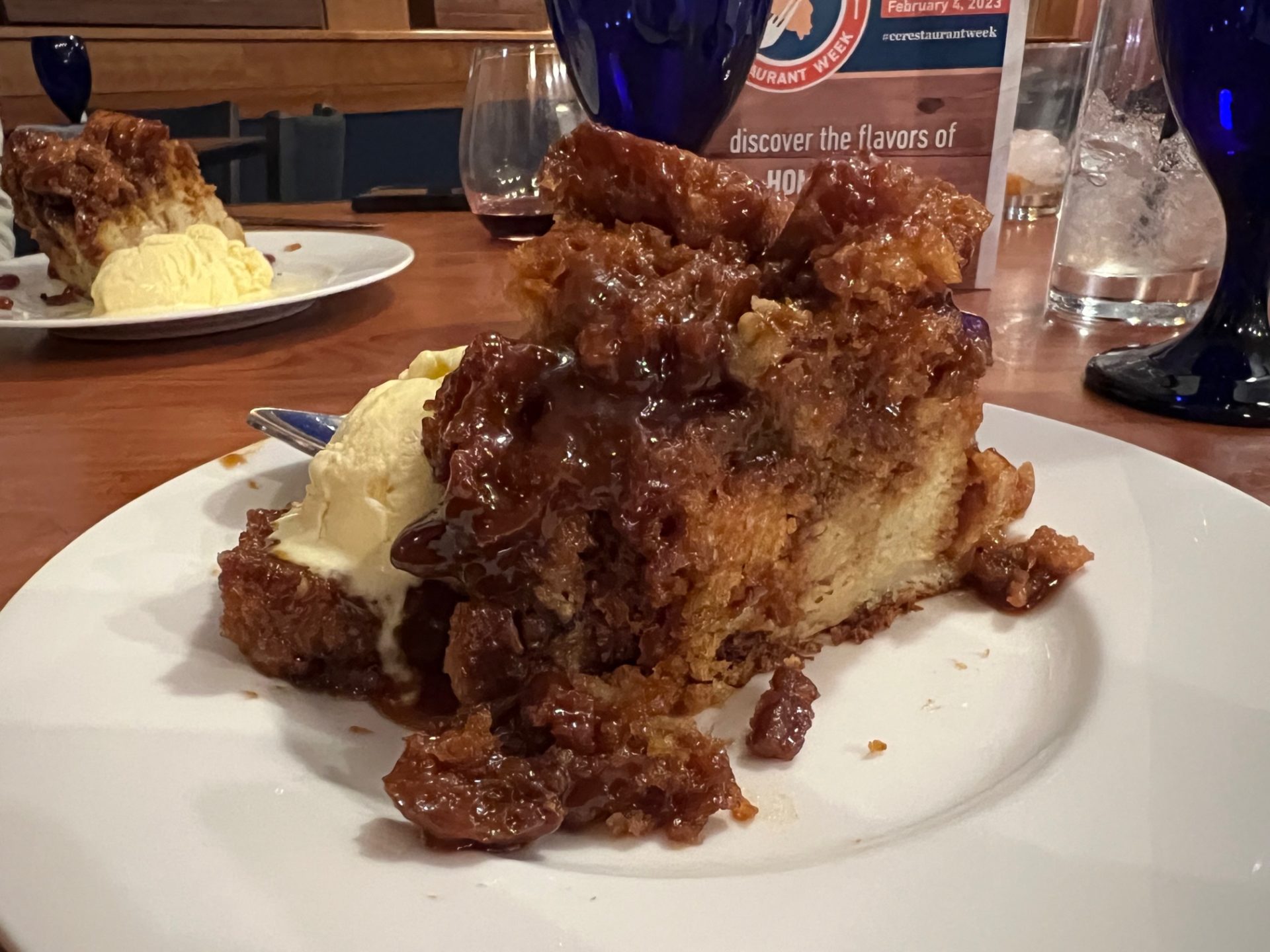 A large slice of bread pudding, covered in a dark brown, thick sauce. Two scoops of vanilla ice cream sit next to it on the round, white plate.