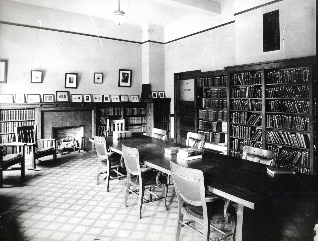 A black and white photo of an old library. It's carpeted and has a long table with several chairs, and bookshelves along each wall.