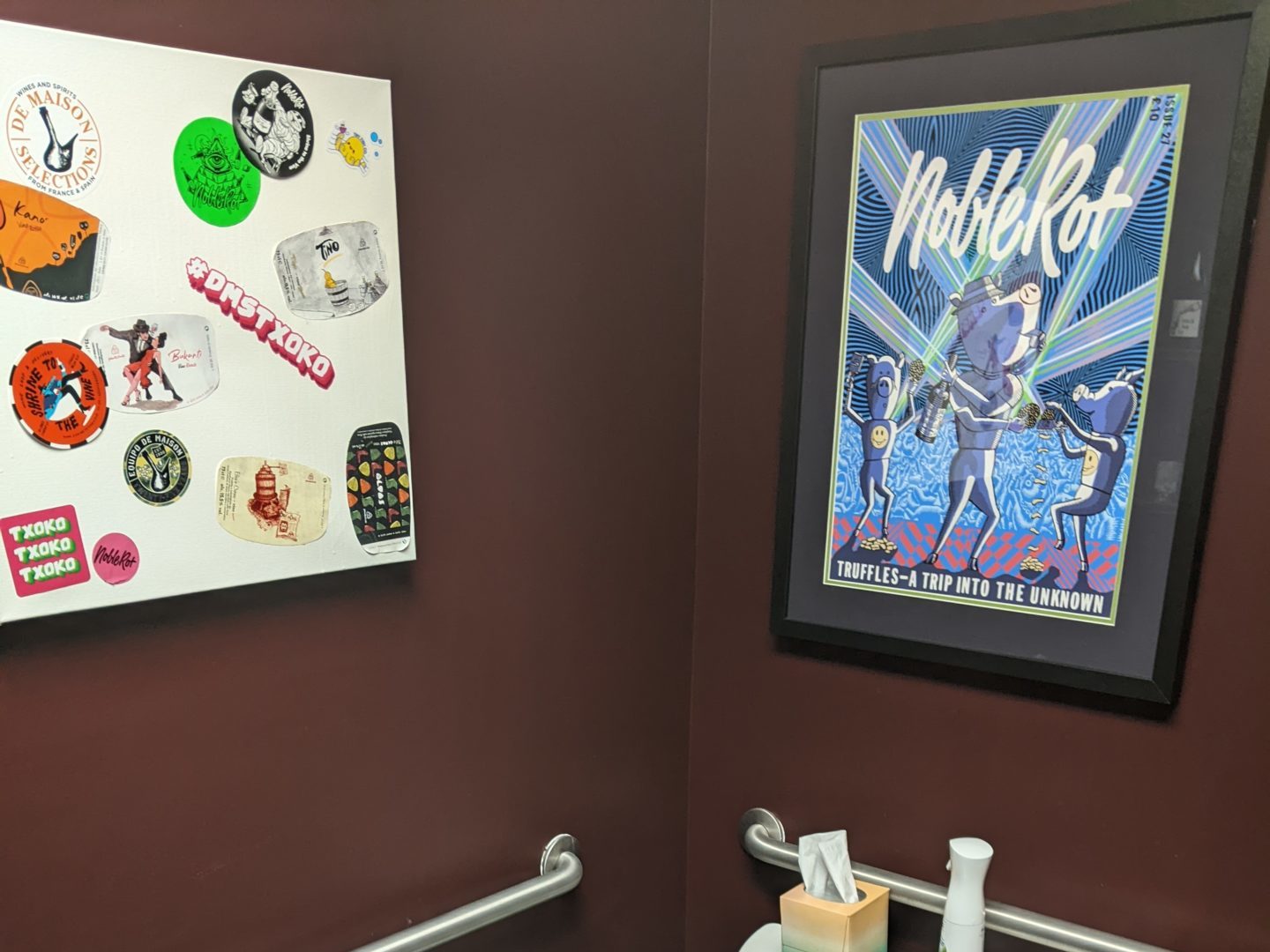 Two wall hangings above a toilet. One is a white board covered in stickers. The other is artwork in a black frame with the words Noble Rot over images of pigs drinking wine.