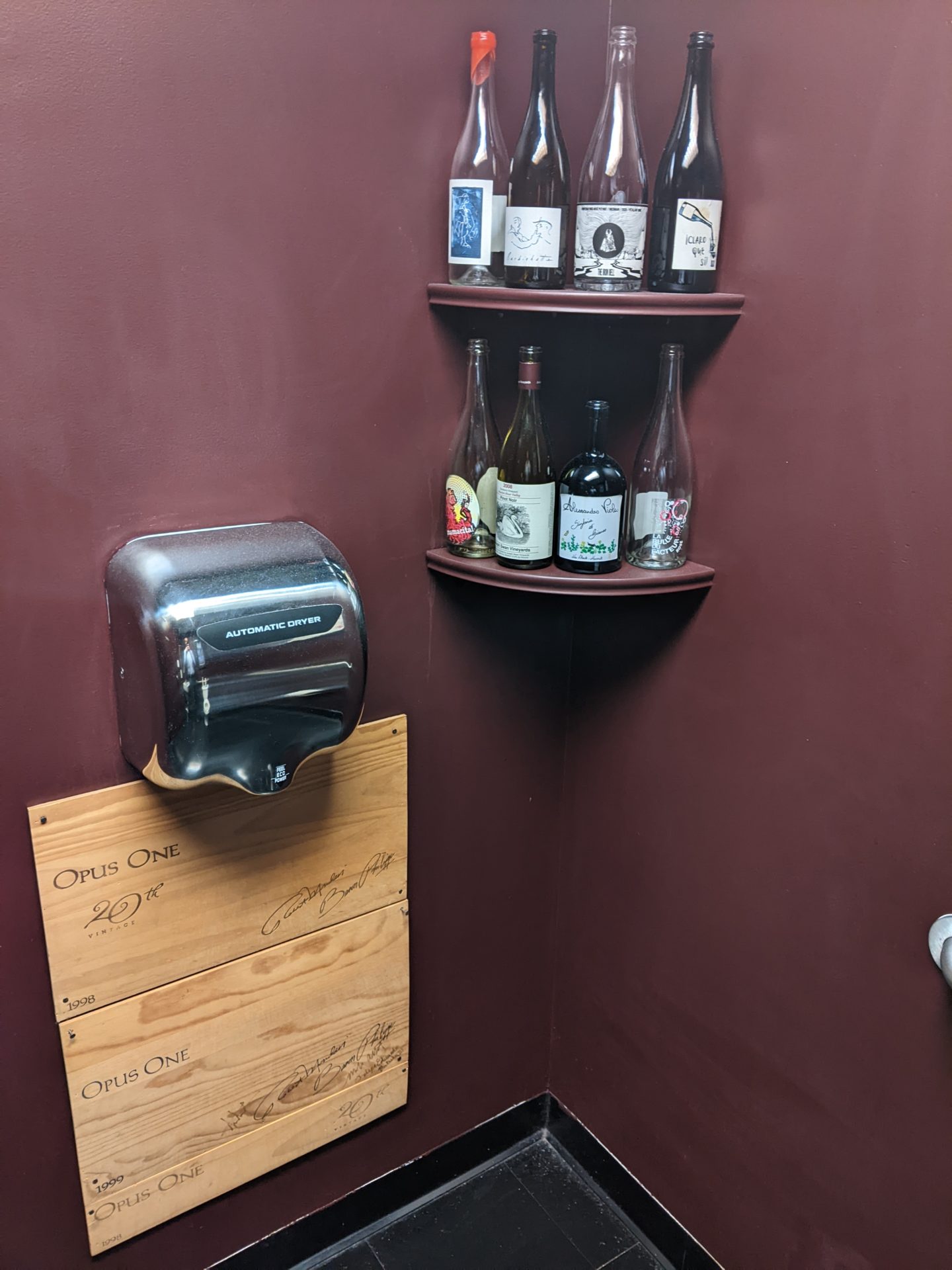 The corner of a room with dark burgundy walls. There are two corner shelves that are filled with empty wine bottles. On one wall there is a stainless steel hand dryer, with two wooden panels attached the wall below it.