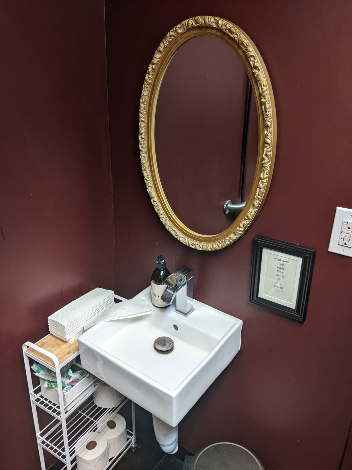 A small square white sink is in the corner of a small room with dark burgundy walls. There is an oval mirror with a gold frame hanging over it. There is a rolling cart with shelves adjacent to the sink.