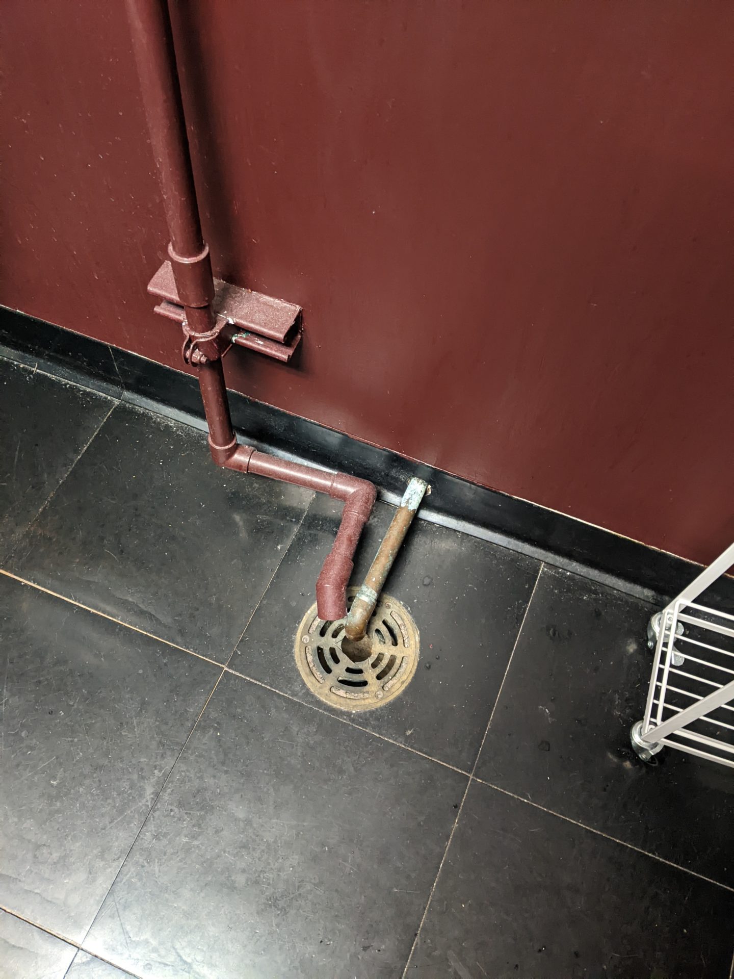A circular drain is built into a black tiled floor. Two pipes lead into the drain.