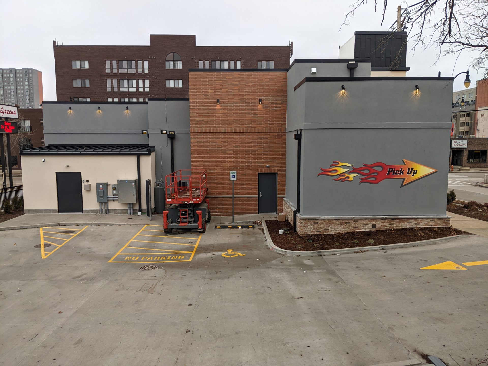 The back side of a building that has sections of gray as well as a section of brick. There are a few parking space in the adjacent lot. On one gray wall is an arrow painted to look like flames that says Pick Up in black lettering.