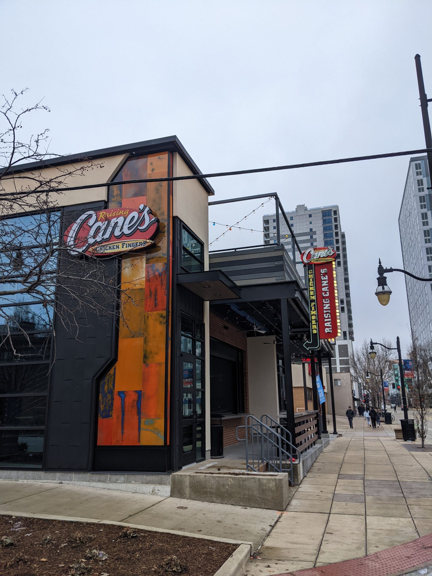 The corner of a building. On one side there's a strip painted in different blocks of color, and a red oval sign that says Raising Cane's Chicken Fingers. On the other side is a marquee style sign that's lit along the side. It's red, and says Raising Cane's.
