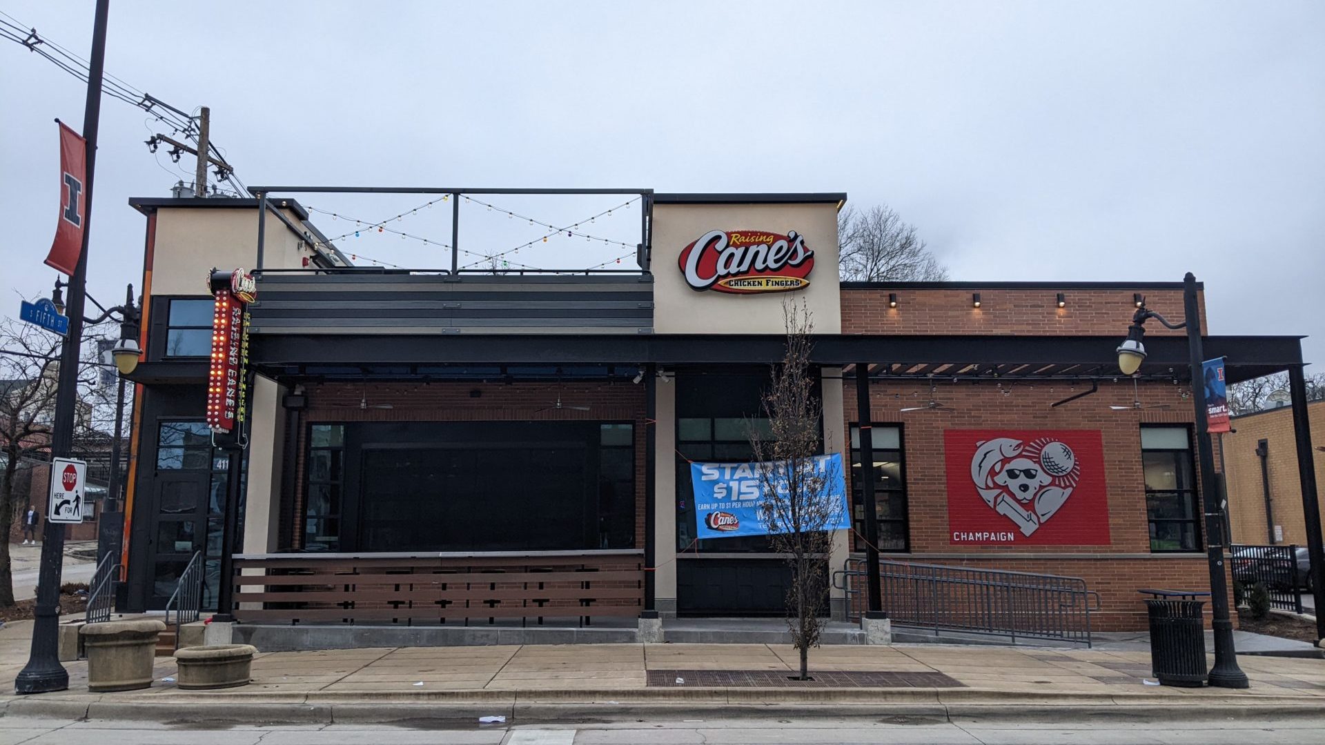 The front of a brick building. It has metal accents and lights strung across the top. There is a sign that says Raising Cane's Chicken Fingers, as well as a square sign with a red background and silver heart comprised of pictures of a dog, disco ball, fish, hat, and block I.