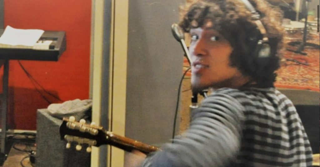 Jose Gobbo holding a guitar looking over his left shoulder, wearing headphones