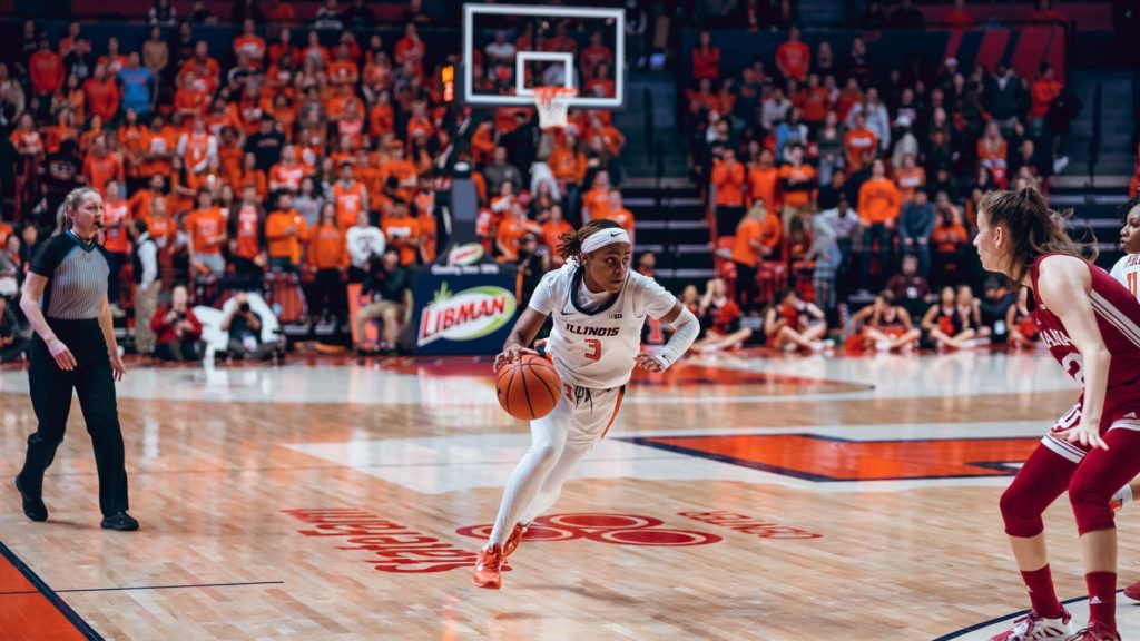 A mid-game action shot of a NCAA women's basketball game. On the court, Illinois women's basketball player Makira Cook is dribbling the ball and moving toward the basket. She is wearing a white jersey (number three) and white headband. Her shoes are orange. The basketball is in her right hand. 