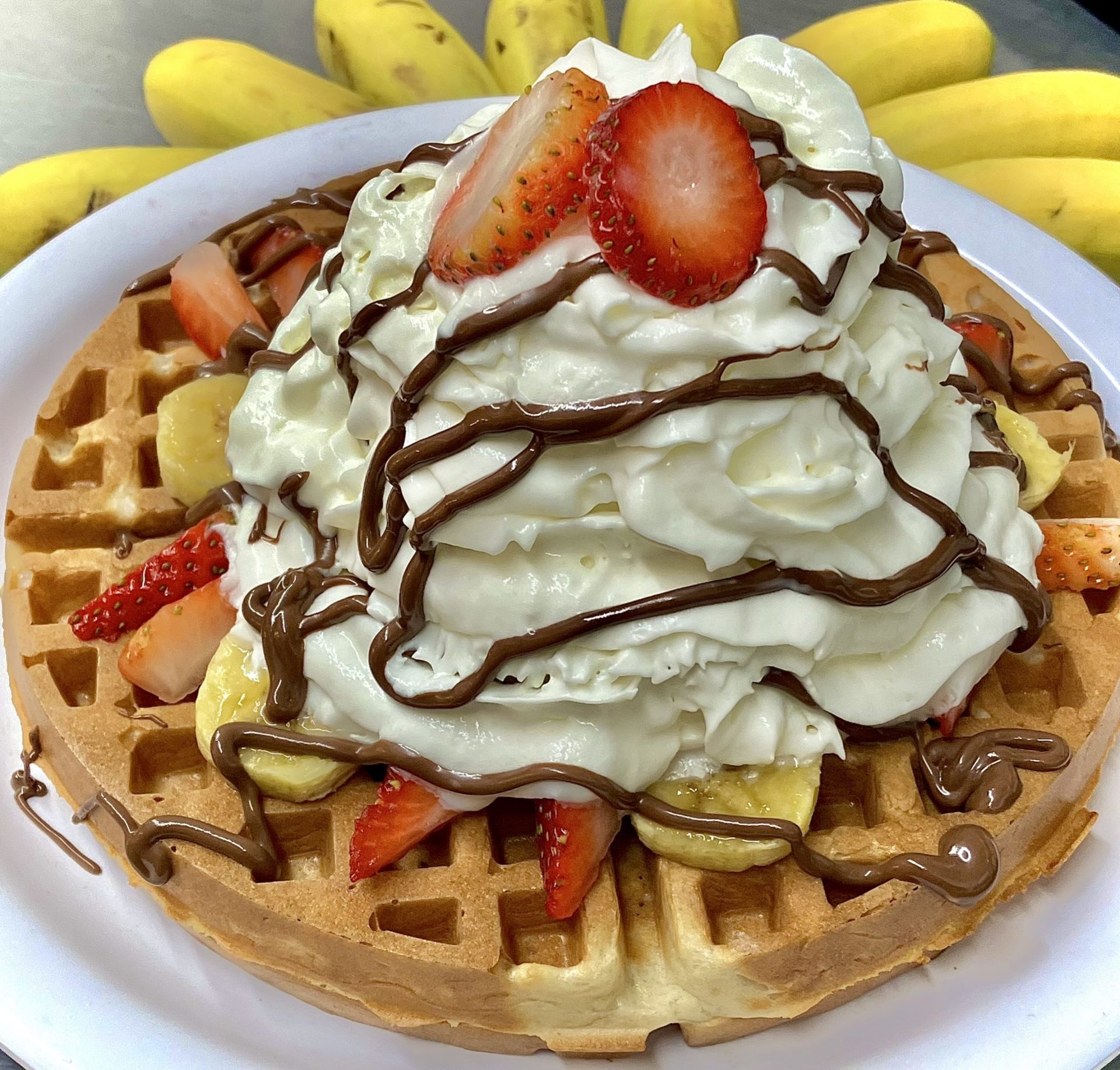 A large waffle topped with sliced bananas and strawberries, a big dollop of whipped cream, and chocolate drizzle.