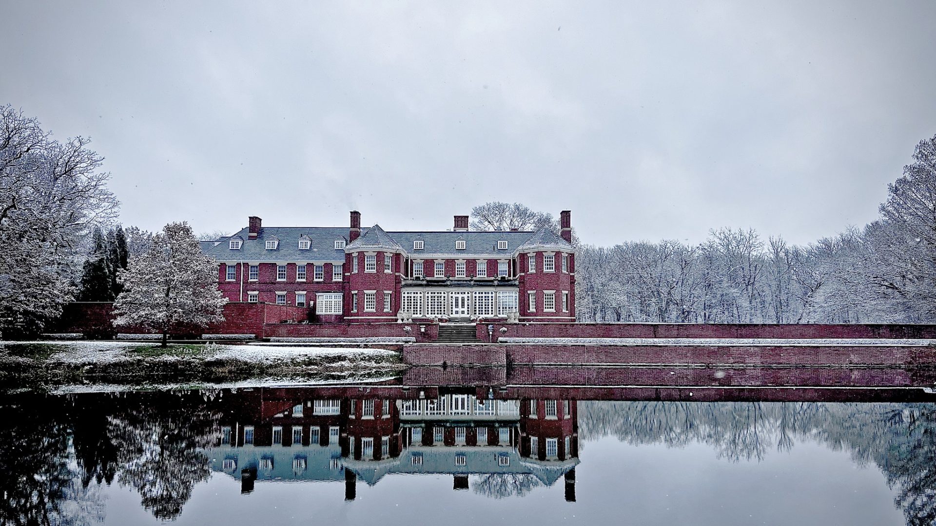 The Allerton Mansion: Red brick with two rows of rectangular windows. In the background are snow covered trees. In the foreground is a pond where you can see the mansion and trees reflected.