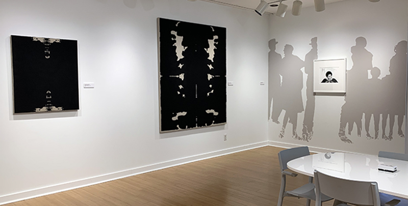 Installation view of Patrick Earl Hammie's artwork. Two paintings with black paint and some linen showing through are on the left and on right is a wall painting of silhouettes of figures over which is a square drawing of a Black woman. In the lower right image of the photo is a table with three gray chairs. 