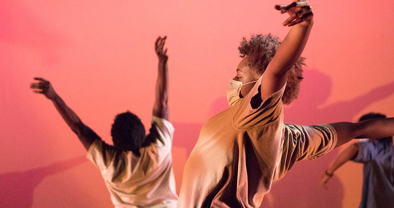 Photo of three dancers in muted beige and blue shirts and masked, raising their arms, each faced in a different direction against a pink wall which shows their shadows.