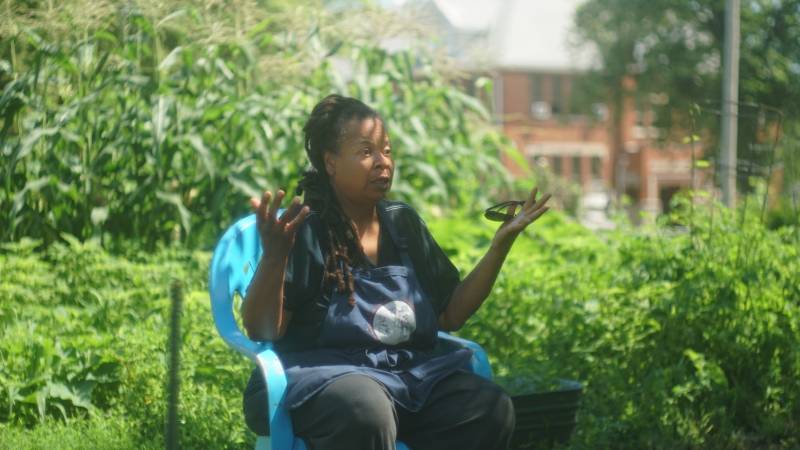 A Black woman with long braids is sitting in a light blue plastic chair, with greenery behind her. She is wearing a black t-shirt and pants, and is gesturing with her hands.