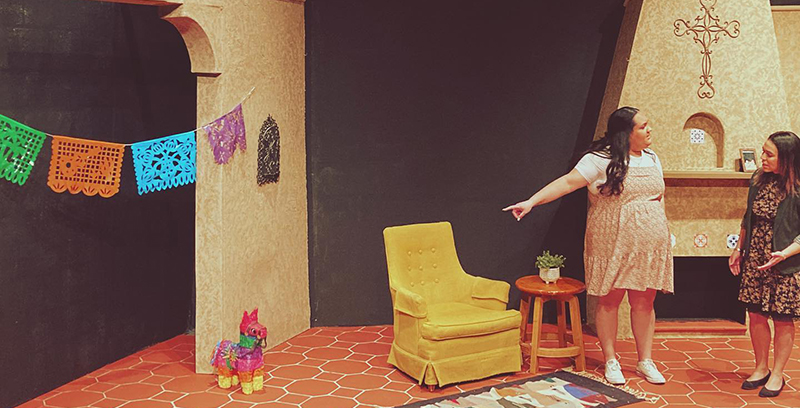 Two female actors arguing off to the left inside a living room with a tall fireplace to the right and archway to the left. Room also contains a yellow easy chair, a colorful rug with a succulent plant on a small wooden stool and a colorful handcut flags hung across the archway and a rainbow pinata on the floor.