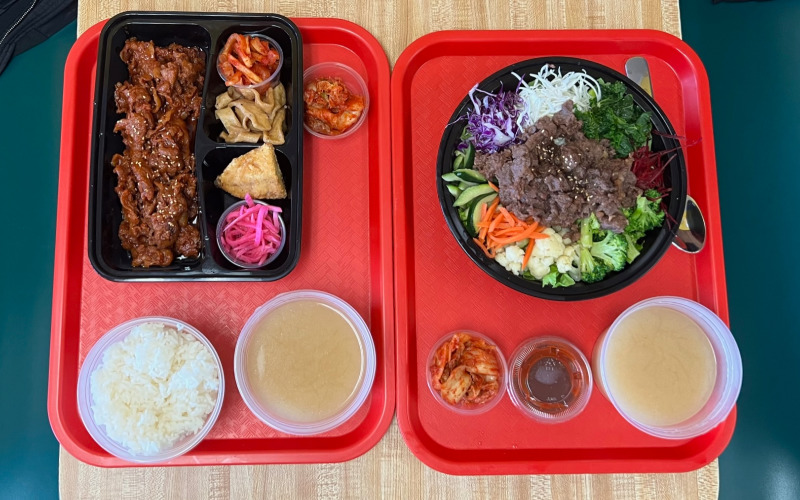 On two red trays, the author's lunch is seen from above. On the left, spicy pork bulgogi with several banchan plus a side of white rice and soup. The right has beef bulgogi bibimbap with a side of kimchi, a spoon, and soup. Photo by Alyssa Buckley.