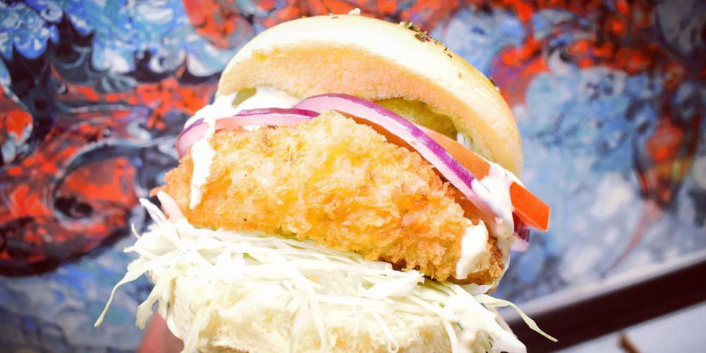 A close, cropped photo of Pond Street's fish sandwich with red onion, pickle, white cabbage, on top of panko catfish. The sandwich is held in front of a red and blue mural. Photo by Pond Street instagram.