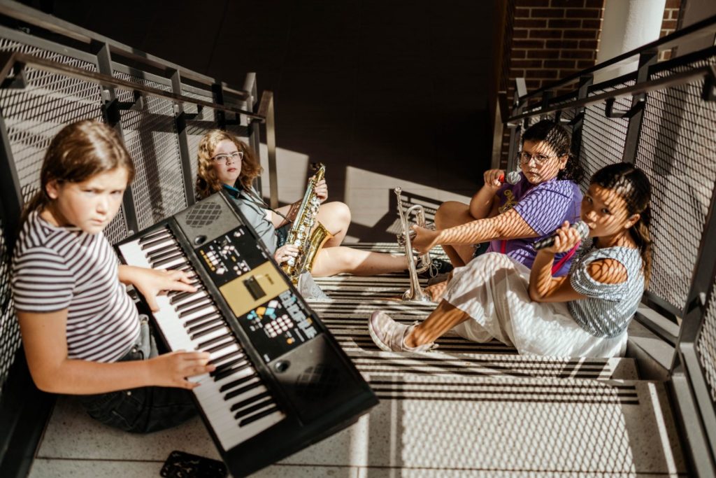 Four girls are sitting on a set of stairs with black railings alongside. One has a large keyboard, one has a saxophone, one is holding a microphone and trumpet, and one has a microphone at her lips.