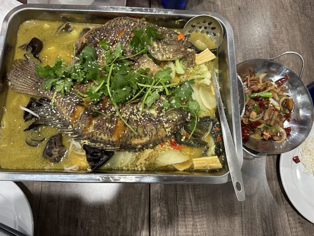 An overhead photo shows two grilled fish in a yellow broth with a metal ladle. Photo by Xiaohui Zhang.