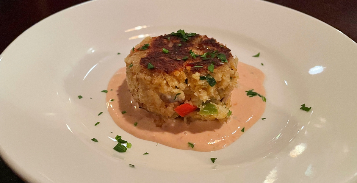A single crab cake sits in the center of a white plate, on a spread of light orange sauce.