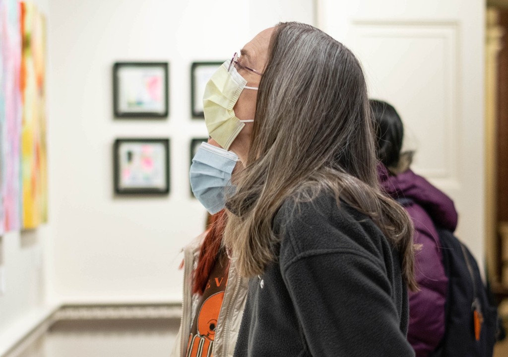 Profile of a woman with long, grayish hair looking at artwork on a gallery wall. She is wearing a dark hoodie and a surgical mask. There are others standing alongside her viewing the artwork as well.