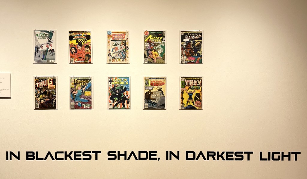 A gallery wall has 10 rectangular art pieces in two rows of five. They look like comic book covers. Along the bottom of the wall it says In Blackest Shade, In Darkest Light in futuristic font.
