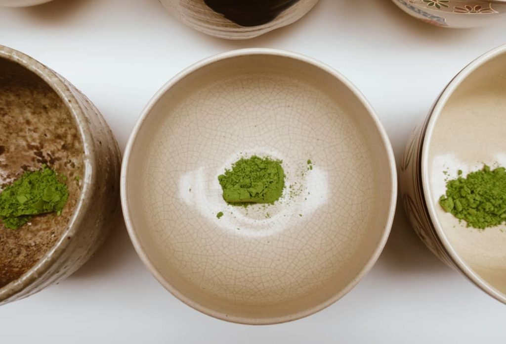 View from above of a beige ceramic bowl with a small amount of green matcha powder in the bottom. It is surrounded by other similar bowls.