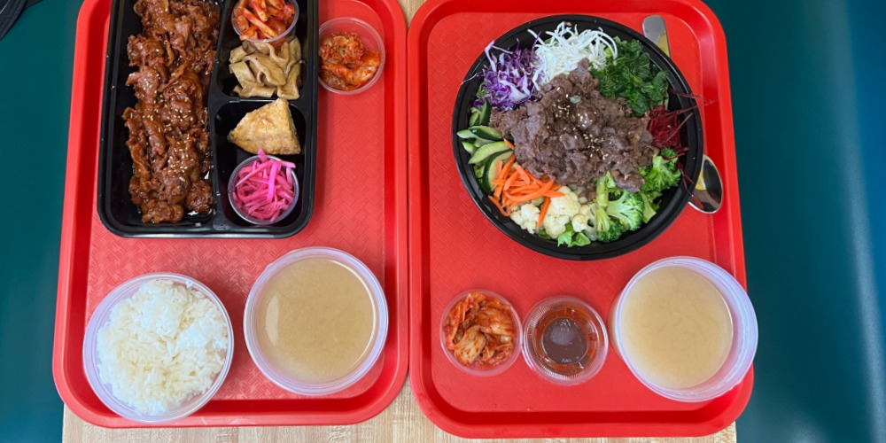 A cropped overhead photo shows the author's meal: spicy pork dosirak and beef bulgogi bibimbap on bright red trays on a teal table at Kimchi Factory in Champaign, Illinois. Photo by Alyssa Buckley.