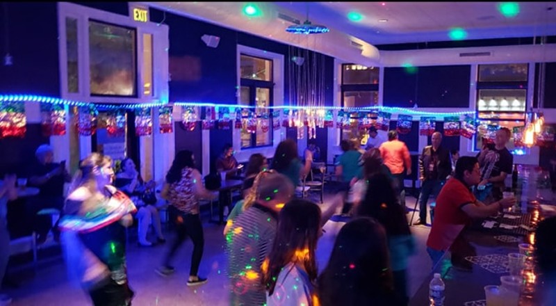 People are scattered across a dark dance floor lit with colored lights. There are also people sitting at a bar.