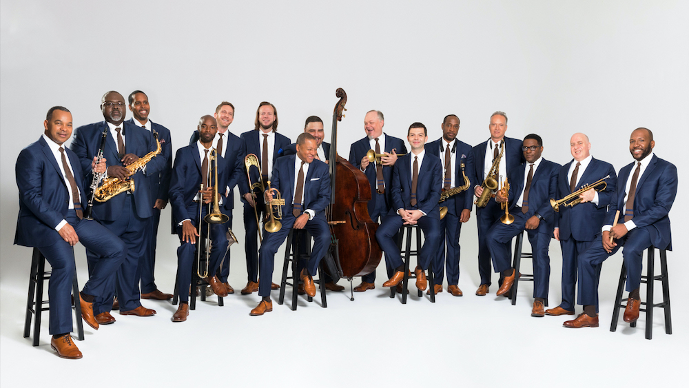 The Jazz at Lincoln Center Orchestra sits in front of a white backdrop, each member holding their respective instruments.