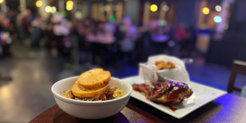 Two Restaurant Week meals at Neil St Blues in Champaign, Illinois sit on a high top table. In the blurred background, the restaurant is busy with full tables and purple party lights. Photo by Alyssa Buckley.
