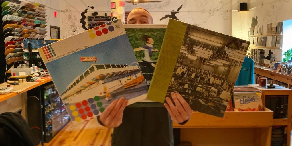 A man is holding three record jackets in front of his face. He is standing in the foreground of a store with wooden floors.