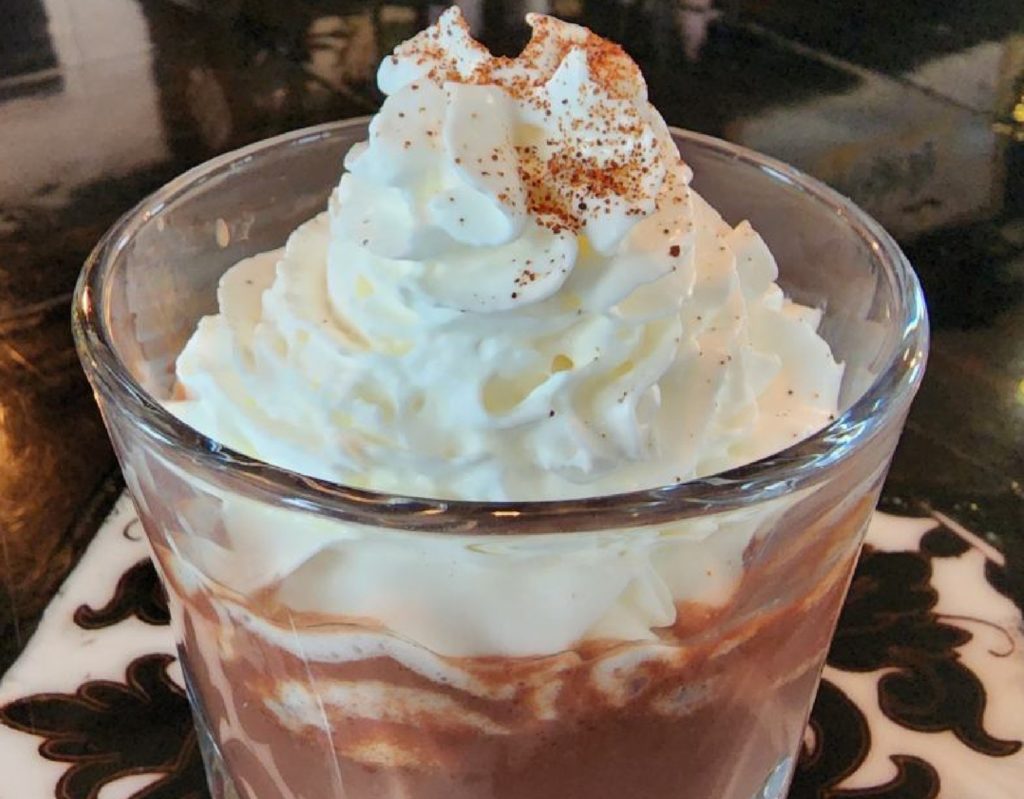 A clear rocks glass is filled with hot chocolate and topped with whipped cream. The whipped cream is sprinkled with cocoa powder.