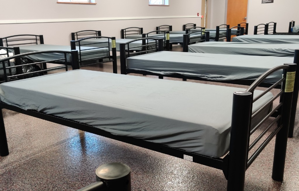 Two rows of cots with gray fitted sheets in a large, bright room.