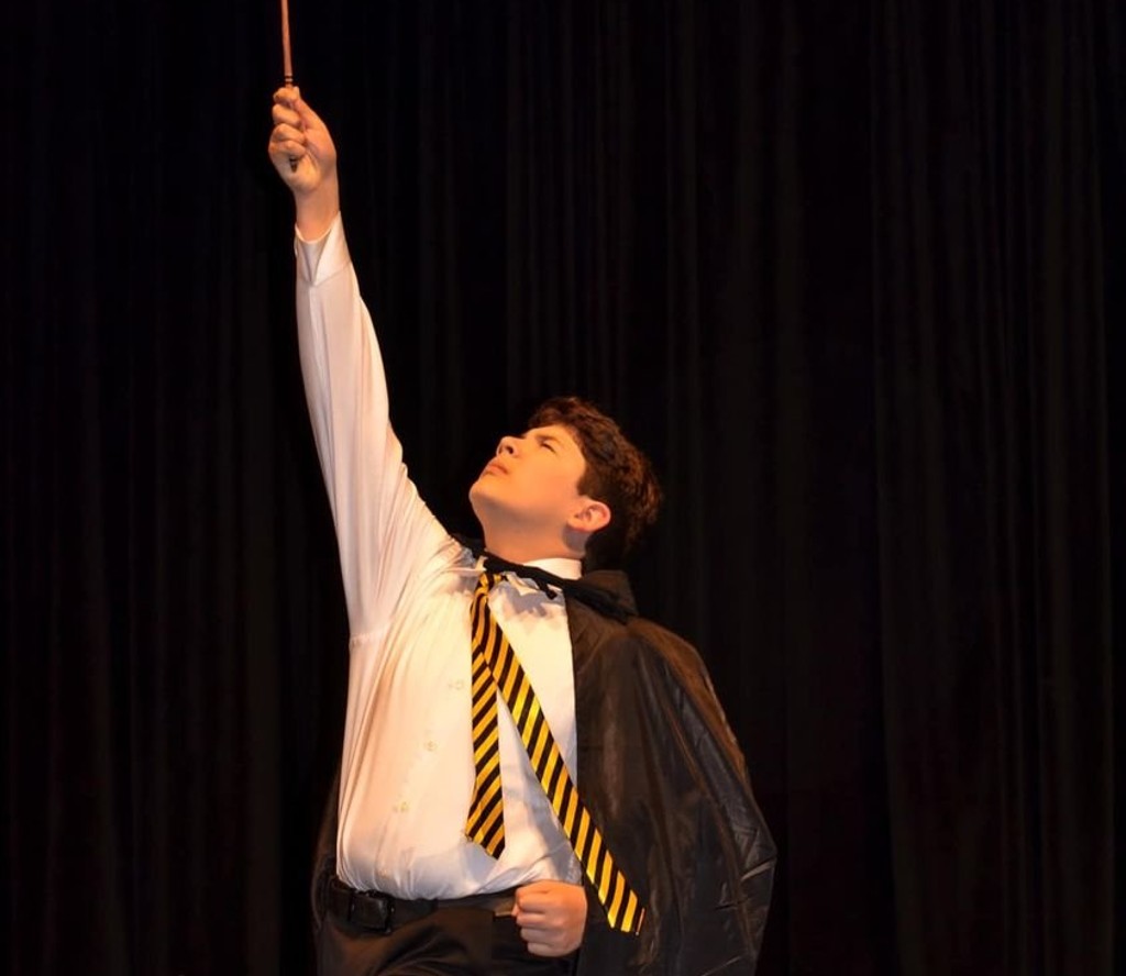 A young man in a white shirt, maroon and gold striped tie, and black cloak is pointing a wand to the sky.