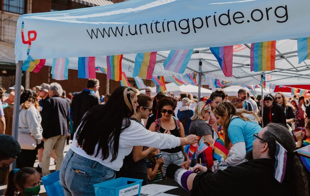 A crowd of people are gathered around a table with a white tent over it. On the edge of the tent it says www.unitingpride.org, and there are various LGBTQ+ flags hanging along the edge.