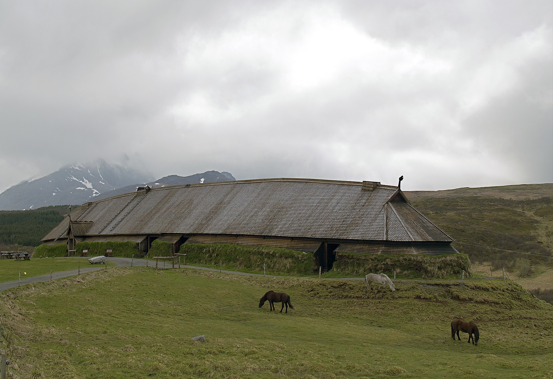 A viking long house: A long wooden structure with a sloped roof. It's in pasture with horses grazing nearby.