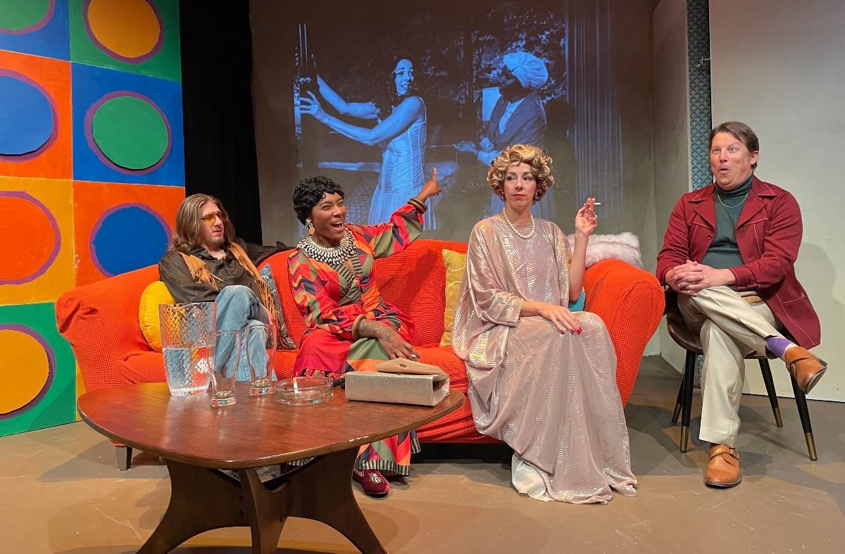 Scene from a play at the Station Theatre. Four people sit on a couch on a set that looks like it is meant to be in the 1970s. The wall on the right is painted in bold colorful circles. On an orange couch from left to right are a white man dressed as a hippie, a Black woman in a 70s-style dress laughing and pointing at a screen behind the couch, (which shows a movie scene with a white woman and a Black woman mammy figure), and a white woman dressed in an elegant gown holding a cigarette. To her right is a white man sittin gin a chair, dressed in a burgundy blazer and making a face of delighted shock.