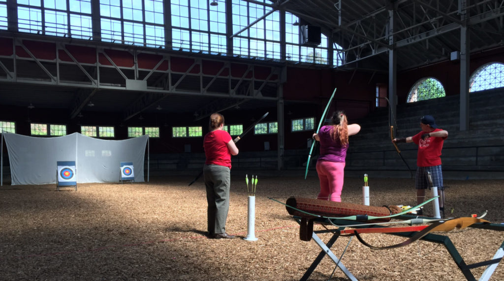 Three people are in the University of Illinois Stock Pavilion, practicing archery. They are standing next to each other and aiming or shooting their arrows at targets a distance away. There is a stand with archery gear in the bottom right of the photo.  