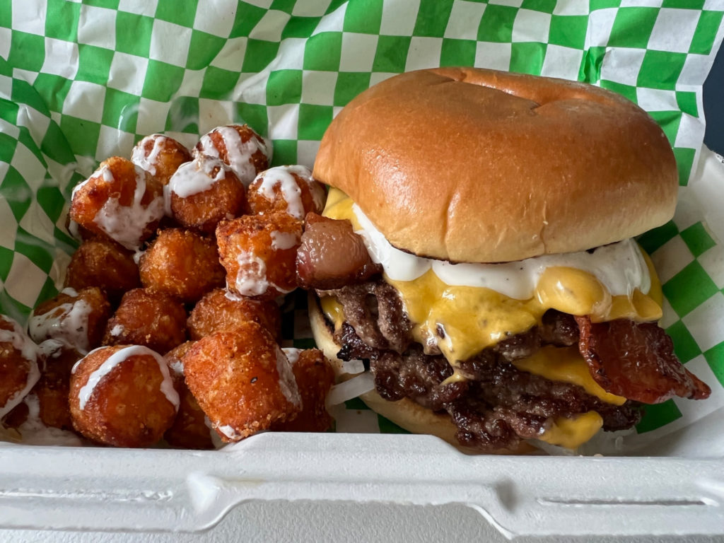 A side of sweet potato tots are drizzled with white marshmallow sauce beside a burger with American cheese and ranch and slices of bacon. The author's order is from Smith Burger Co food truck and is inside a styrofoam container lined with green-and-white parchment paper. Photo by Alyssa Buckley.