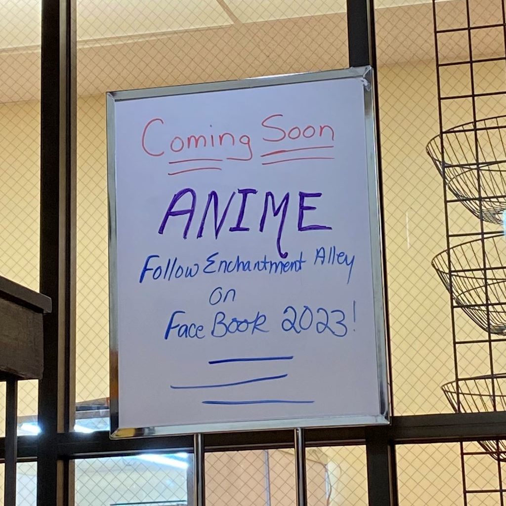 A rectangular dry erase board is hanging in a window. It says "Coming Soon, ANIME. Follow Enchantment Alley on Facebook 2023"