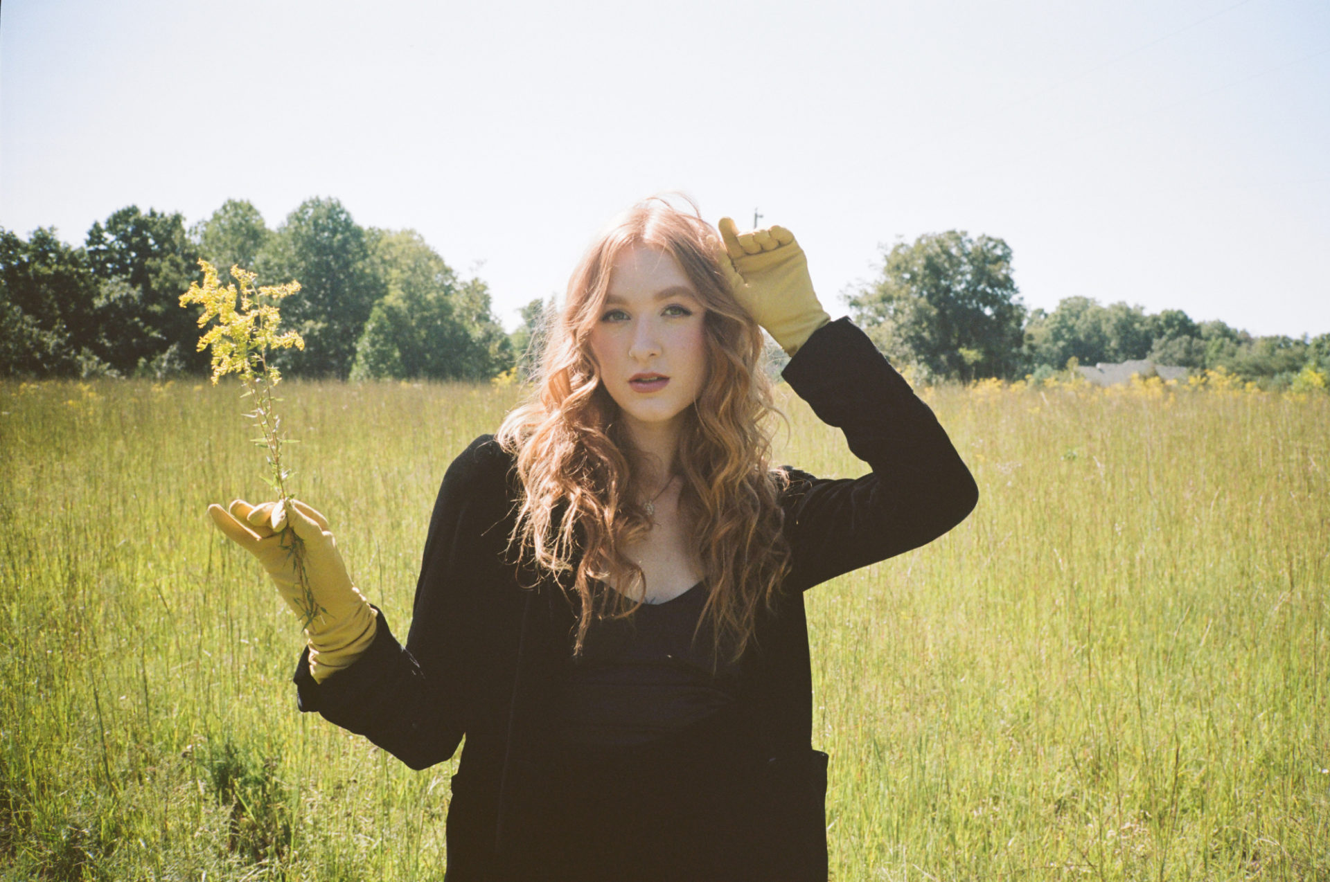 Photo of Bella White. A thin, white woman with long, wavy reddish hair is standing in a field of tall grass. She is wearing all black, including a black jacket, and chartreuse gloves that match the color of the grass behind her. Her left arm is raised to the side of her head and he rright arm holds a yellow flower. Photo by Bree Fish.