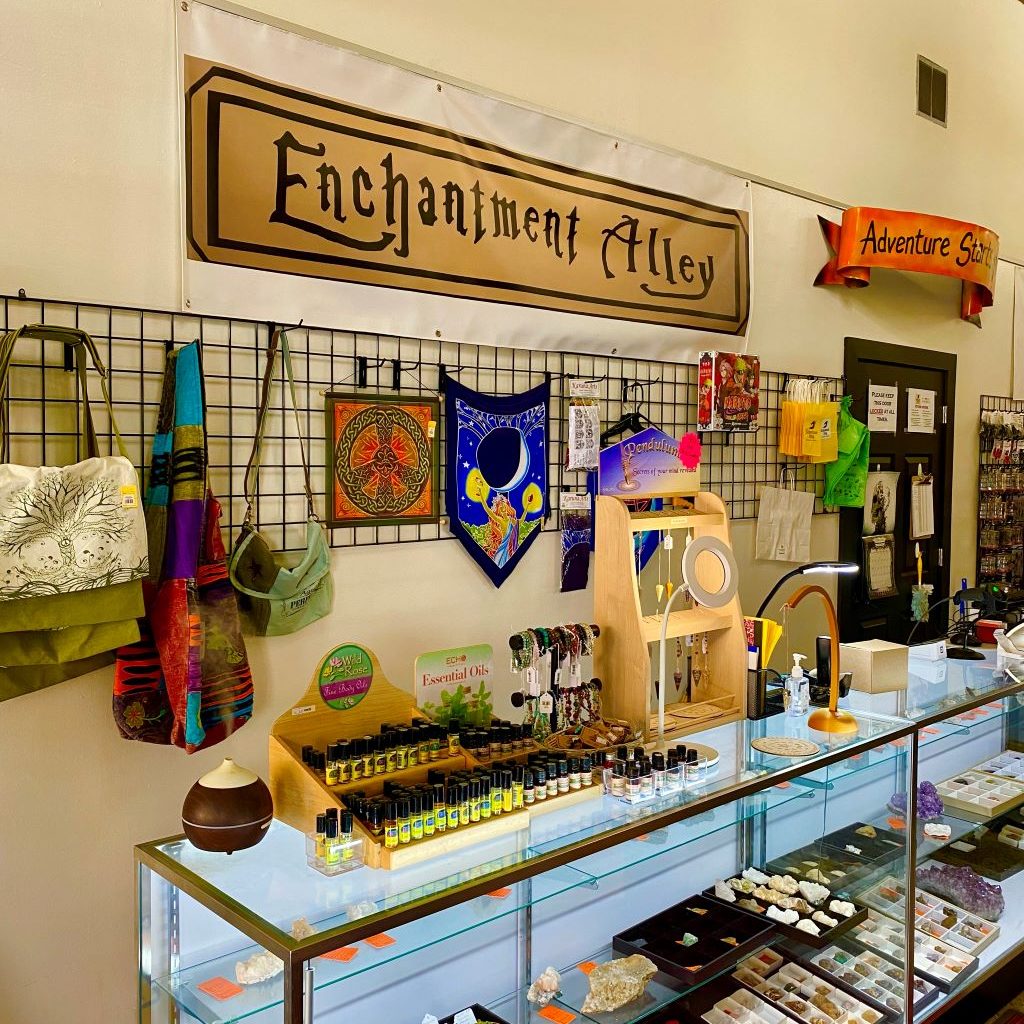 A glass case is filled with trays of various crystals. On top of the case there are displays of oils and pendants. Behind the case is a rack hanging on a wall with small tapestries and handbags hanging on it. Above the rack is a gold sign with Enchantment Alley in black lettering.