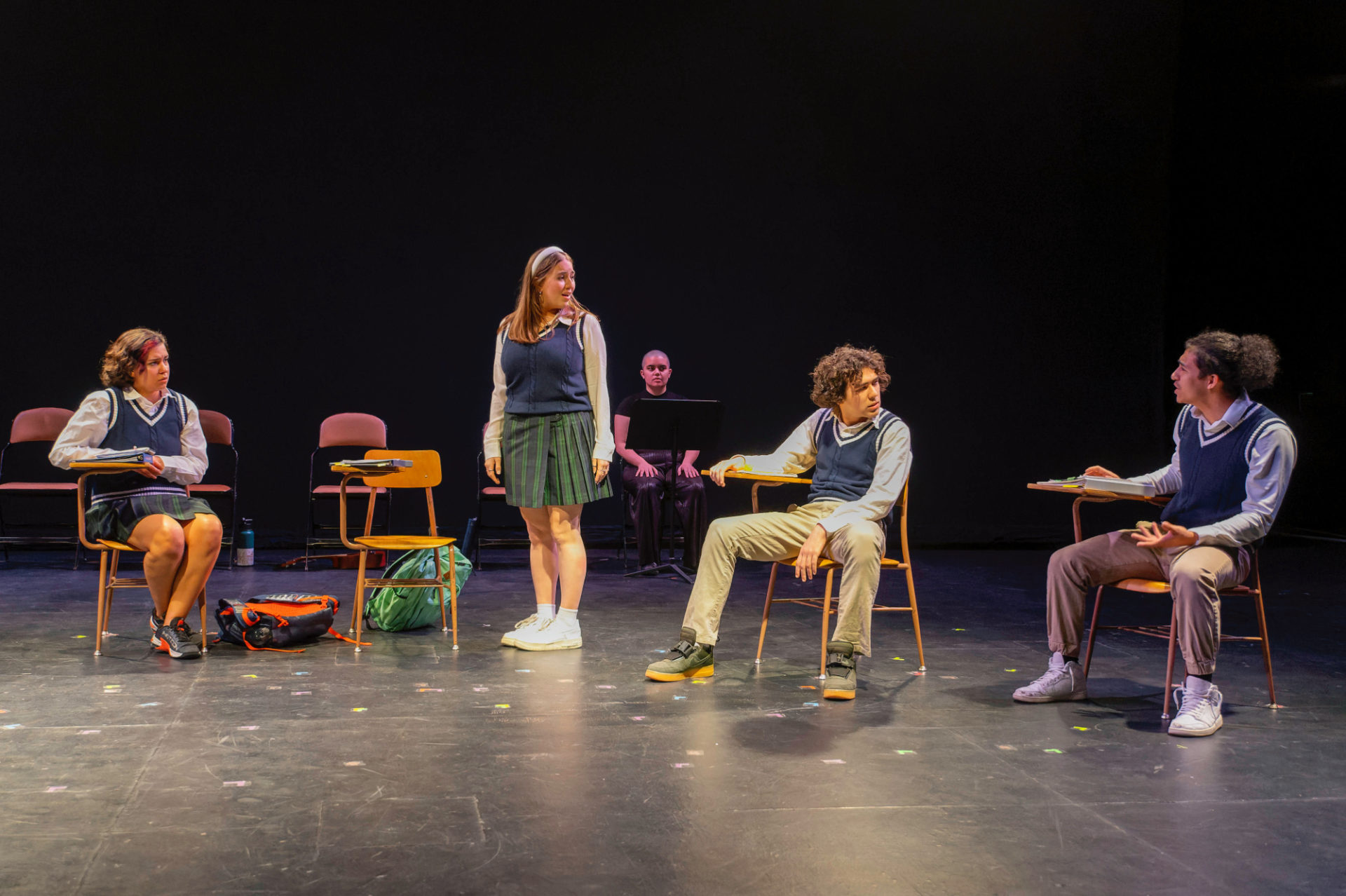 Four young people are on a theatre stage. They are all wearing school uniforms. Three are sitting down, one woman is standing up. There is a person in black sitting behind them and watching.