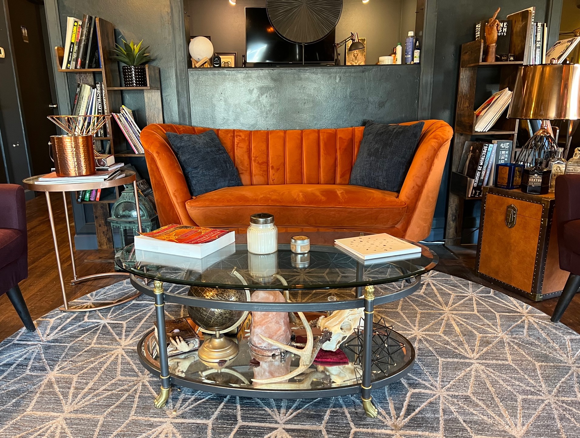 An orange couch sits up against a wall with a window cut out of it. There are bookshelves on either side, and a glass table in front, sitting on a patterned rug. 