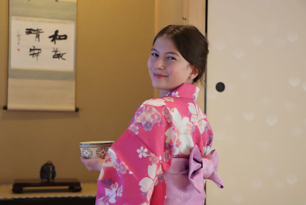 A girl in a pink flowered yukata is looking over her shoulder at the camera. She is holding a ceramic bowl.