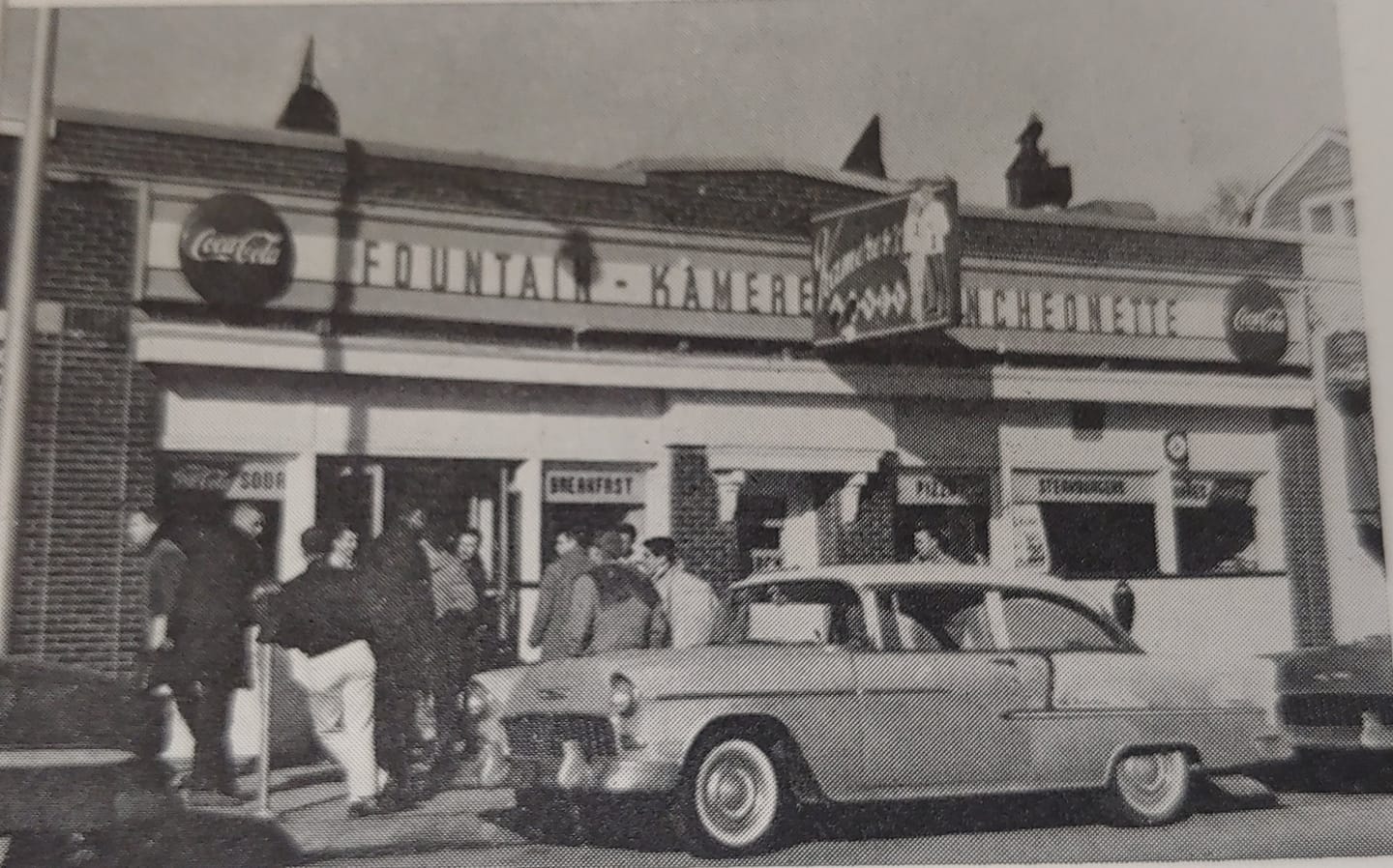 A black and white photo of the front of a luncheonette. There is a 1950s style car parked in front, and several people milling around outside.