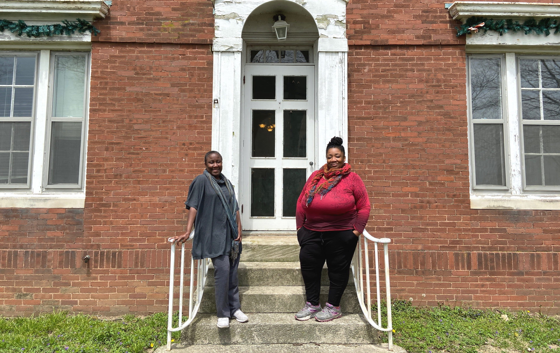 Two Black women are standing on concrete steps, each leaning against a white metal railing. The steps lead to the entrance of a red brick building with a white door.