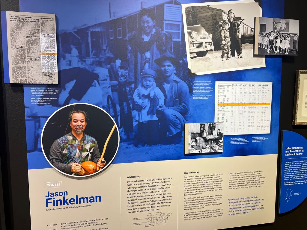 A board with a description of the artwork on display. There is a Picture of Jason Finkleman and a description about his work below. The top of the image features a picture in blue with black and white photos in the corners