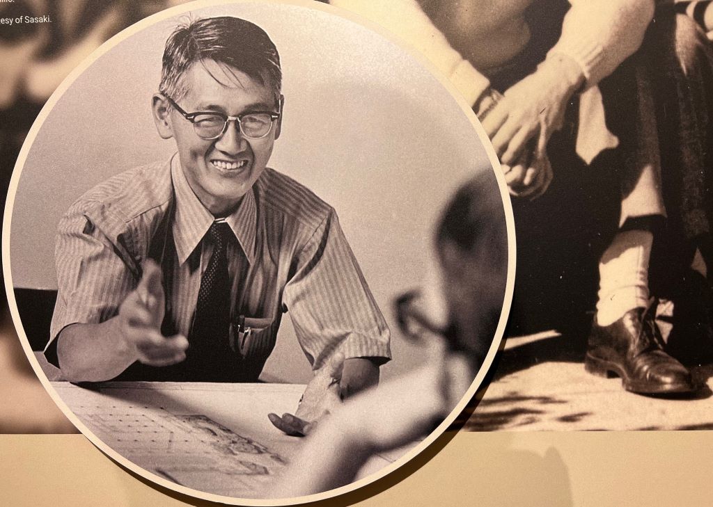Close up of a photo displayed on a museum panel. It's a black and white photo of a Japanese American man in glasses, a striped shirt, and black tie. He is smiling and gesturing to someone across from him.